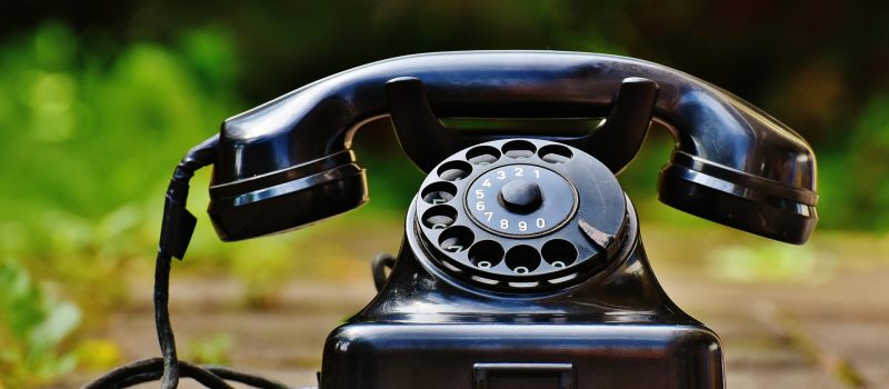 Close-up of a classic black rotary phone outdoors with a blurred green background.